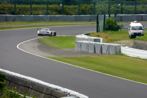 Ein Rennwagen fährt auf einer Rennstrecke mit einem Polizeiwagen im Hintergrund, umgeben von Gras, Pflanzen, Bäumen und einem Fahrzeug auf der rechten Seite.