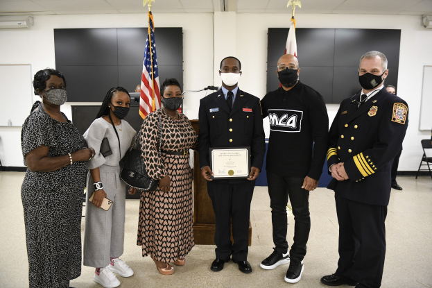 Eine Gruppe maskierter Menschen vor Flaggen, wobei eine Person in der Mitte ein Zertifikat hält, während es scheint, als fände eine Abschlussfeier statt.