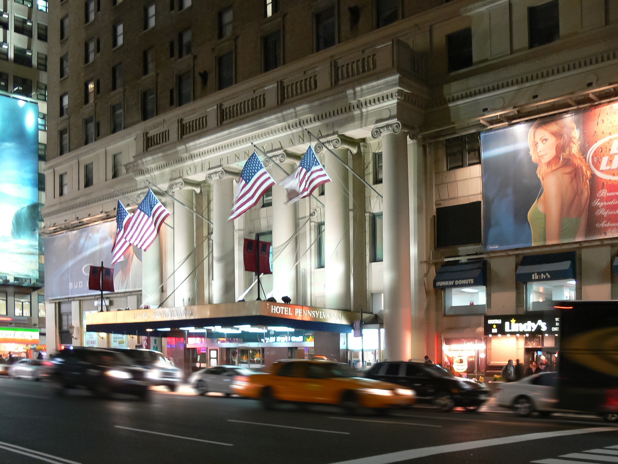 Eine nächtliche Stadtstraße mit fahrenden Autos, ein Gebäude mit Fenstern, Säulen und Fahnenmasten, beleuchtete Schilder mit Bildern und Text sowie helles Straßenlicht.