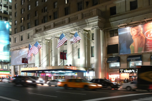 Eine nächtliche Stadtstraße mit fahrenden Autos, ein Gebäude mit Fenstern, Säulen und Fahnenmasten, beleuchtete Schilder mit Bildern und Text sowie helles Straßenlicht.