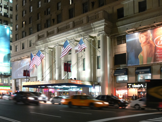 Eine nächtliche Stadtstraße mit fahrenden Autos, ein Gebäude mit Fenstern, Säulen und Fahnenmasten, beleuchtete Schilder mit Bildern und Text sowie helles Straßenlicht.