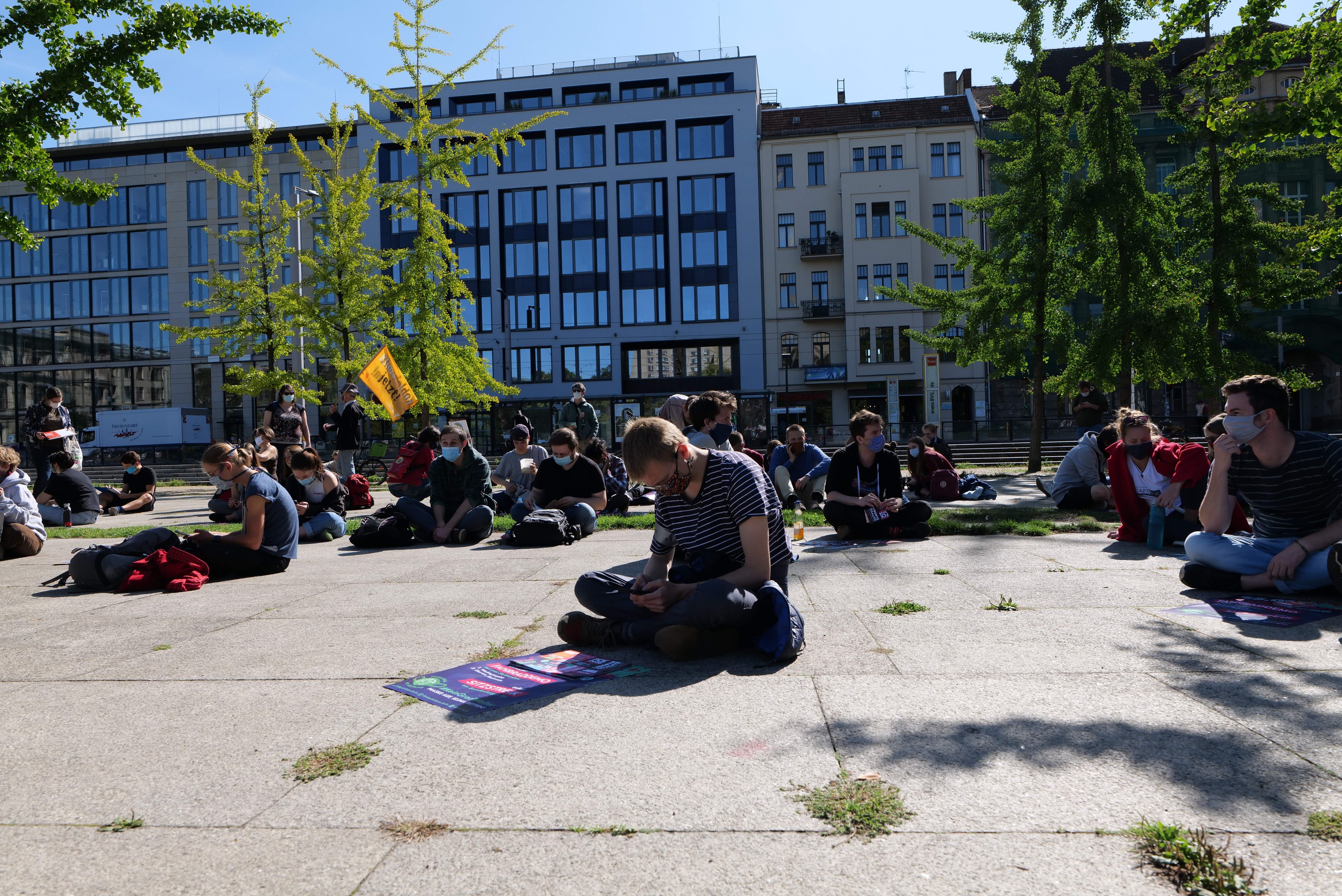 Eine Gruppe von Menschen sitzt vor einem Gebäude auf dem Boden während einer Demonstration in Berlin, einige tragen Masken und sind von verstreuten Taschen und Bäumen umgeben unter einem klaren blauen Himmel.
