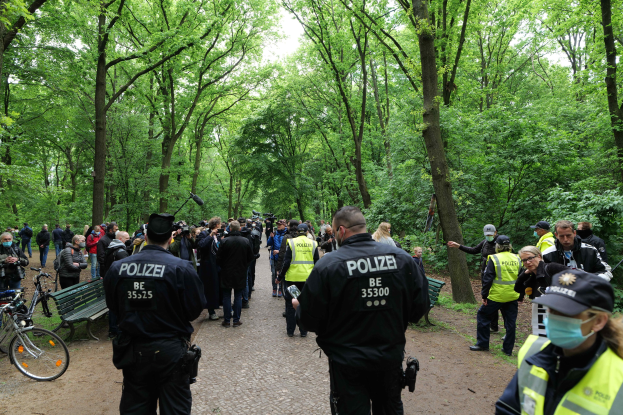 Polizeibeamte vor einer Menschenmenge bei einer Anti-Terror-Demonstration in Berlin, mit Fahrrädern und einer Bank im Vordergrund und Bäumen im Hintergrund.