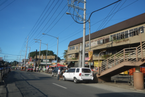 Eine Stadtstraße mit Autos, Fußgängern, Gebäuden, Strommasten, Laternen, Bannern, Bäumen und einem Himmel im Hintergrund.