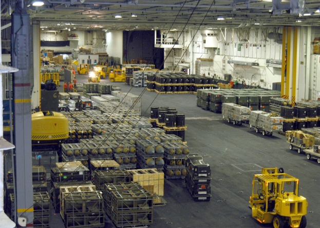 Großes Lagerhaus-Innere mit mehreren Lastwagen, Containern, Menschen, Beleuchtungsausrüstung, Metallstangen und einem sichtbaren Rad.