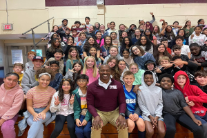 Schüler posieren für ein Foto in einer Turnhalle bei einem Sommercamp, wobei einige auf der Treppe sitzen und andere stehen, eine Tür und ein Ausgangsschild auf der linken Seite sichtbar