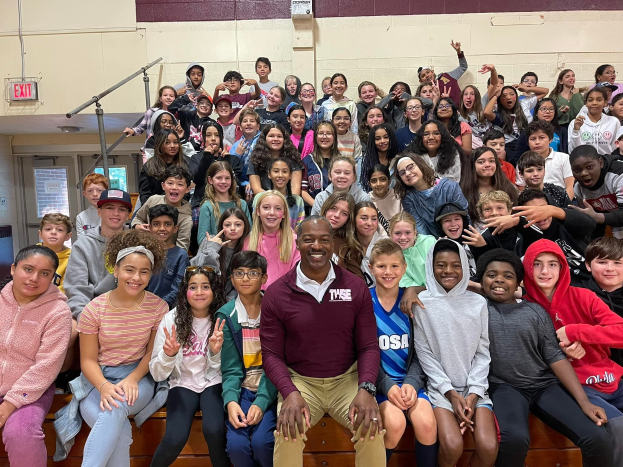 Schüler posieren für ein Foto in einer Turnhalle bei einem Sommercamp, wobei einige auf der Treppe sitzen und andere stehen, eine Tür und ein Ausgangsschild auf der linken Seite sichtbar