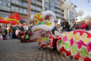 Eine Löwen-Tanzvorstellung während eines Chinesischen Neujahrsfestes in Amsterdam, mit einer Zuschauermenge und Menschen, die den Moment mit Kameras festhalten, vor einer Kulisse aus Gebäuden und einem klaren blauen Himmel.