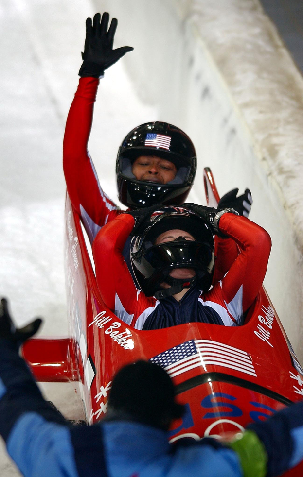 Zwei Bobsleighs auf einer Bahn, einer mit sichtbarem Helm und Handschuhen, mit einer Person unten und einer Wand im Hintergrund.