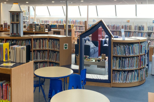 Eine Bibliothek mit Bücherregalen, Tischen, Stühlen, Sofas mit Kissen, einem Pfahl, Deckenlampen und anderen Gegenständen.