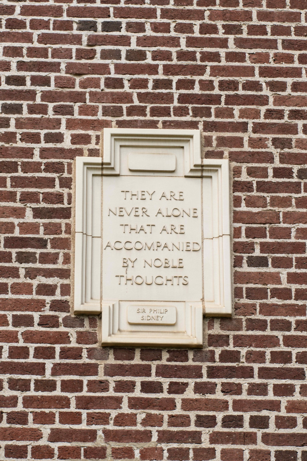 Eine Backsteinmauer mit einer darauf angebrachten Steinplatte, die den Spruch "Sie sind nie allein, die von edlen Gedanken begleitet werden" in fetter Schrift zeigt.