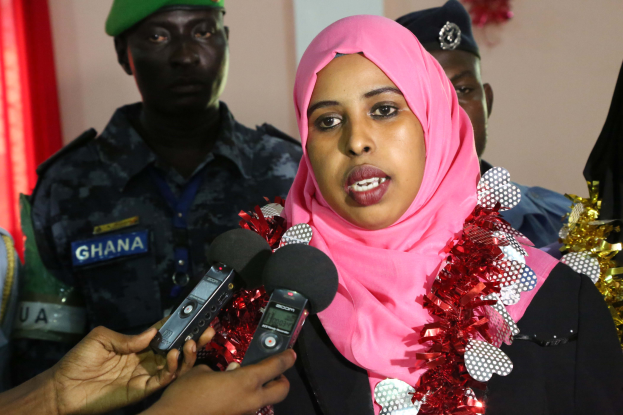 Eine Frau mit pinkem Kopftuch spricht zu Reportern, mit einem Mikrofon von der linken Seite des Bildes gehalten, vor einer Wand mit einem Vorhang im Hintergrund.