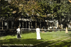 Eine Gruppe von Menschen, die Tennis auf einem Rasenplatz vor dem Abenakis Springs Hotel in Provo, Quebec, spielen, wobei einige Rackets halten, andere auf Stühlen sitzen, ein Netz in der Mitte und Bäume und ein Gebäude im Hintergrund, mit Text am unteren Bildrand.