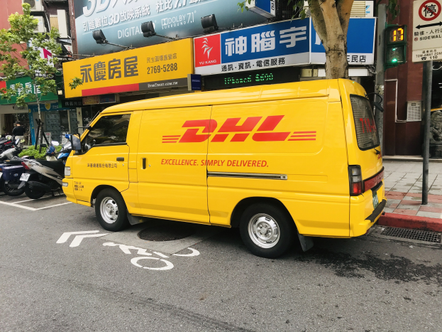 Gelber DHL-Lieferwagen auf der Straße geparkt, umgeben von Gebäuden, Bäumen, Texttafeln, Pfählen, Verkehrsschildern und einer Person auf dem Gehweg.