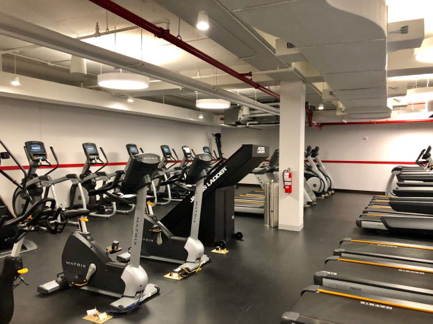 Ein Fitnessstudio mit mehreren Laufbändern und Ellipsogertäten in einer Reihe, einer zentralen Säule, einem wandmontierten Feuerlöscher und Deckenleuchten in einem Fitnessstudio in New York City.