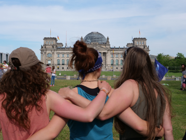Drei Frauen in bunten Kleidern stehen vor dem Reichstaggebäude in Berlin, mit einer Menge, Fahnen und Bäumen im Hintergrund.