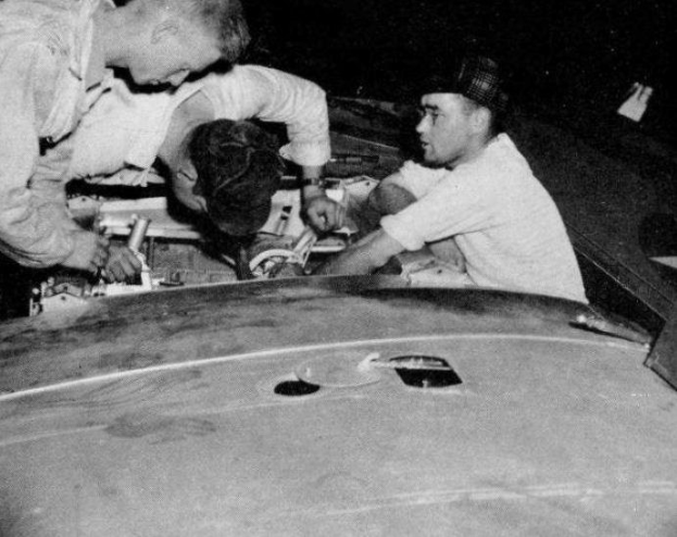Drei Männer in Kappen reparieren ein schwarz-weißes Auto in einer Garage umgeben von Werkzeugen und Materialien.