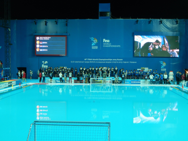 Eine Gruppe von Menschen um einen Swimmingpool mit einem Netz am Boden herumstehend mit einer Wand im Hintergrund, die "FINA World Championships 2015 Kazan"-Text, Logos, Träger und Lichter anzeigt.