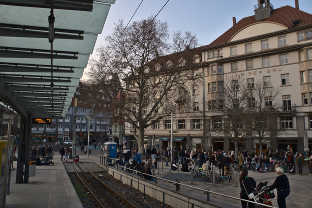 Eine belebte Stadtstraße mit einem Bahnhof auf der linken Seite, umgeben von Gebäuden, Bäumen und Laternen, mit Menschen, die gehen, sitzen und stehen, in der Nähe des Bahnhofs unter einem sichtbaren Himmel.