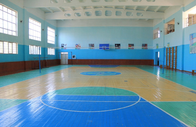 Große Turnhalle mit einem blauen und weißen Basketballfeld, einem Netz an Stangen, einer Tür, Fenstern, Plakaten an den Wänden, einem Geländer und einem Dach.