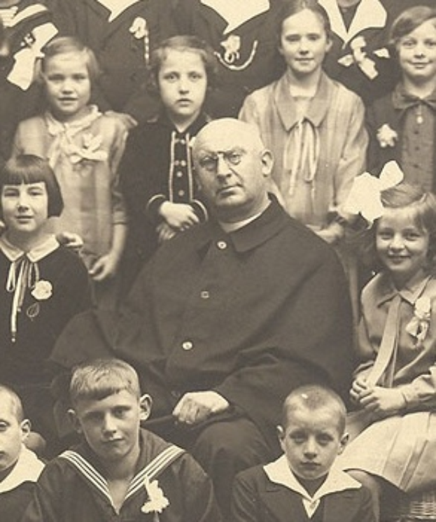 Ein altes Schwarz-Wei├č-Foto eines ernst aussehenden Mannes, der in der Mitte sitzt und von einer Gruppe Kinder umgeben ist, die ihn bewundernd anschauen.