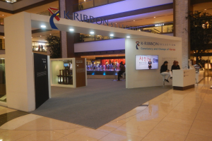 Ein großer Ausstellungsstand in einem Einkaufszentrum mit Menschen, die darauf sitzen und stehen, mit Texttafeln, Lichtern, Säulen, Pflanzen und einem sichtbaren Boden.