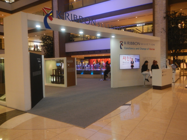 Ein großer Ausstellungsstand in einem Einkaufszentrum mit Menschen, die darauf sitzen und stehen, mit Texttafeln, Lichtern, Säulen, Pflanzen und einem sichtbaren Boden.