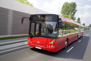 Ein roter Bus fährt eine Straße entlang neben einem Gebäude, mit einer Person im Inneren, neben einer Wand, Bäumen, Laternen und einem bewölkten Himmel.