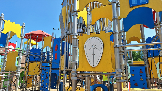 Ein Spielplatz mit buntem Spielgerät, einschließlich Stangen, Pfählen und einem Schirm, einige Menschen, Bäume und einen klaren blauen Himmel.
