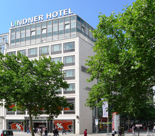 Großes Gebäude mit Glasfront und "Lindner Hotel"-Schriftzug oben, umgeben von Bäumen und einer belebten Straße mit Fußgängern und Fahrzeugen unter einem sichtbaren Himmel.