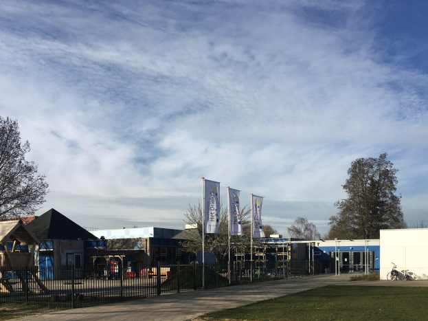Großes Gebäude mit einem grasbewachsenen Bereich davor, umgeben von einem Metallzaun, mit einem Weg, Bäumen, Pfosten, Bannern, einem geparkten Fahrrad, nahen Häusern und einem bewölkten Himmel, als das neue Kinderspielgelände an der Schule identifiziert.