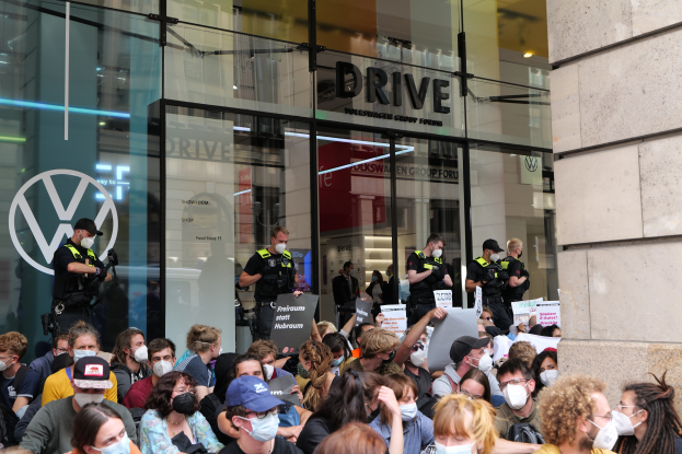 Eine große Gruppe maskierter Demonstranten mit Schildern vor einem Glasfrontgebäude in Melbourne, dokumentiert von Fotografen.