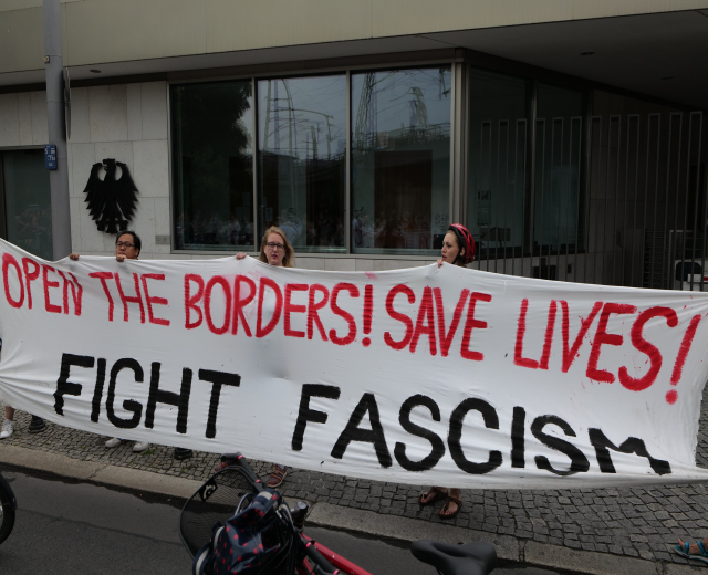 Demonstranten mit einer Transparent mit der Aufschrift "öffnet die Grenzen, rettet Leben, bekämpft Faschismus" vor einem Gebäude mit Glasfenstern und einem Mast, mit einem Fahrrad und einer Tasche auf der Straße.
