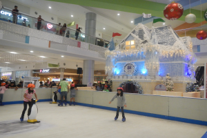 Eine Gruppe von Menschen, die auf einer Eisbahn in einem Einkaufszentrum Schlittschuh laufen, umgeben von einer hauseigenen Struktur, die mit Lichtern und festlichen Dekorationen geschmückt ist, mit Zuschauern und zusätzlichen Dekorationen, die eine festliche Atmosphäre schaffen.