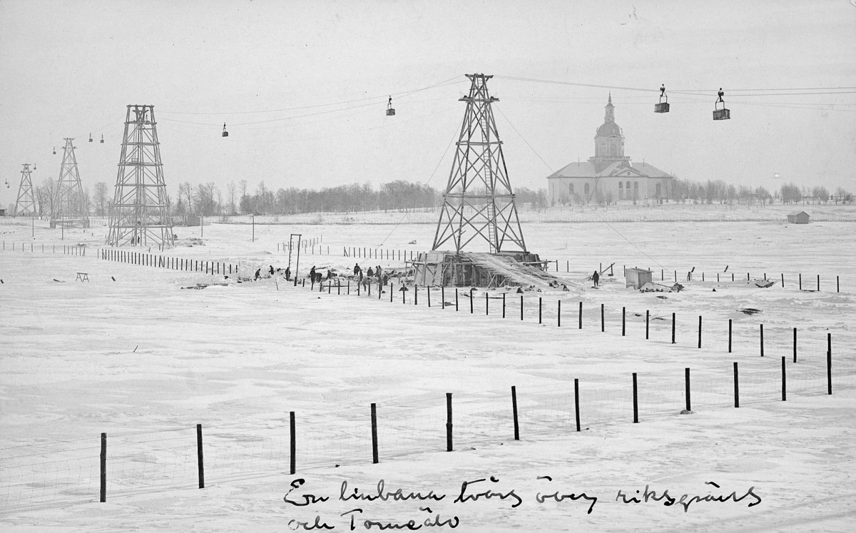 Schwarze-Weiß-Foto eines Skilifts in einem verschneiten Feld mit Stützpfählen, Überseilbahn, Bäumen und einem Gebäude im Hintergrund, mit Text unten.