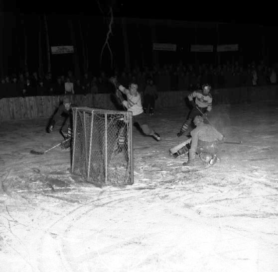 Schwarzes und weißes Foto eines Hockey-Spiels auf einem Eisstadion mit Spielern in der Nähe eines Tors und Zuschauern im Hintergrund.
