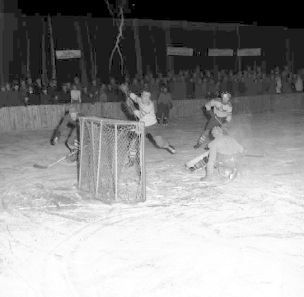 Schwarzes und weißes Foto eines Hockey-Spiels auf einem Eisstadion mit Spielern in der Nähe eines Tors und Zuschauern im Hintergrund.