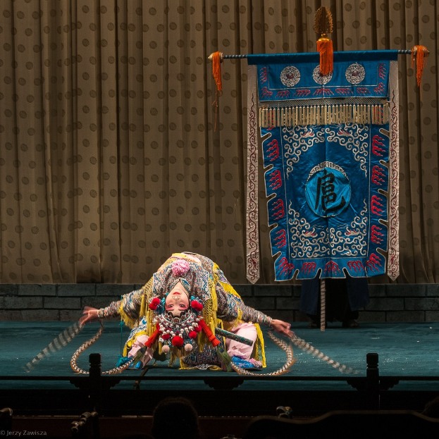 Eine Frau in buntem Kostüm performs chinesische Oper auf einer Bühne mit Vorhang und Banner im Hintergrund, während das Publikum unten zuschaut.