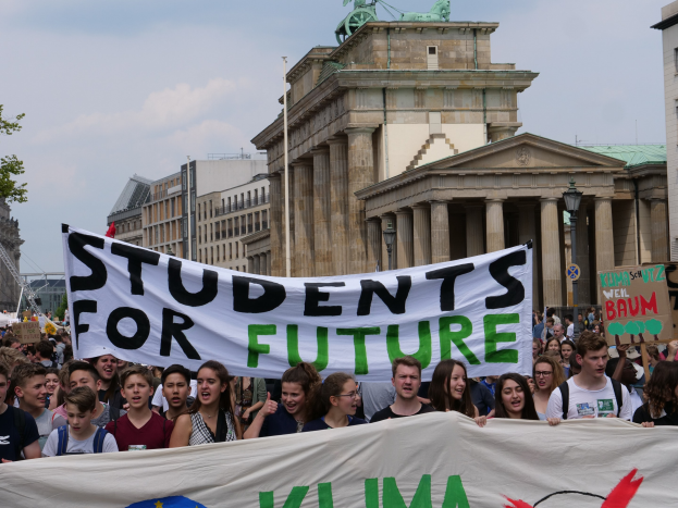 Gruppe von Studenten marschiert in Berlin mit einem buntfarbenen 'Students for Future'-Schild an Gebäuden, Bäumen und Himmel vorbei.
