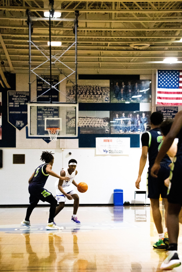 Gruppe junger Männer beim Basketballspielen in einer Turnhalle mit Wand, Deckenstangen und Lampen sowie einer Flagge auf der rechten Seite.