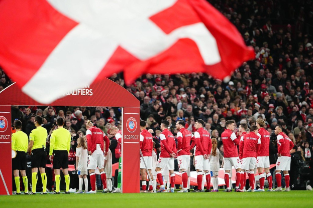 Eine Gruppe von Menschen auf einem Fußballfeld mit einer roten und weißen Fahne im Vordergrund, einem Bogen mit der Aufschrift "Bayern München vs Bayern München Wetten & Vorschau" im Hintergrund und einer großen Menschenmenge im Stadion.
