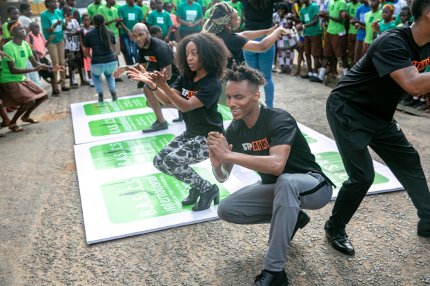 Eine Gruppe von Menschen in grünen Hemden und schwarzen Hosen tanzt auf einer Bühne während einer St. Patrick's Day-Feier, mit einer Menge, die darunter steht und Gebäuden, Bäumen und einem klaren blauen Himmel im Hintergrund.