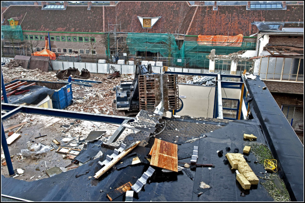 Baustelle mit einem zentralen Kran, umgeben von Gebäuden, Bäumen, Absperrungen und anderen Gegenständen, mit Text am unteren Rand.