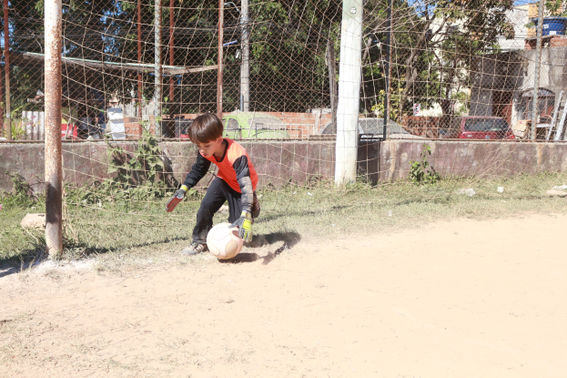 Ein junger Junge spielt Fütball auf einem Schmutzfeld mit einem grasigen Rand, umgeben von Böumen, Pfosten, einem Zaun, einer Wand, Fahrzeugen, Geböuden und einem bewölktem Himmel, trögt Schuhe wöhrend er aktiv im Spiel ist.