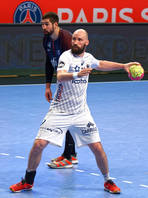 Zwei Männer beim Handballspielen auf einem Platz, einer hält den Ball, mit einer Tafel im Hintergrund, auf der 'Paris Saint-Germain vs Paris Saint-Germain' steht.