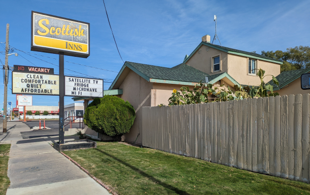 Außenansicht eines schottischen Motels in San Francisco, Kalifornien, mit einem Haus mit Dach und Fenstern, einem Holzzaun, Pflanzen, Gras, Bäumen, Schildern, Strommasten mit Drähten, einer Straßenlaterne, Fahrzeugen auf der Straße und einem bewölkten Himmel.