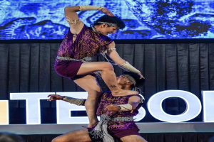 Zwei Akrobaten führen auf der Bühne eine Darbietung auf, wobei der Mann die Frau in seinen Armen hält, während beide purpurfarbene Kleider tragen, mit einer Menge und einem Bildschirm im Hintergrund.