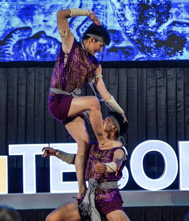 Zwei Akrobaten führen auf der Bühne eine Darbietung auf, wobei der Mann die Frau in seinen Armen hält, während beide purpurfarbene Kleider tragen, mit einer Menge und einem Bildschirm im Hintergrund.