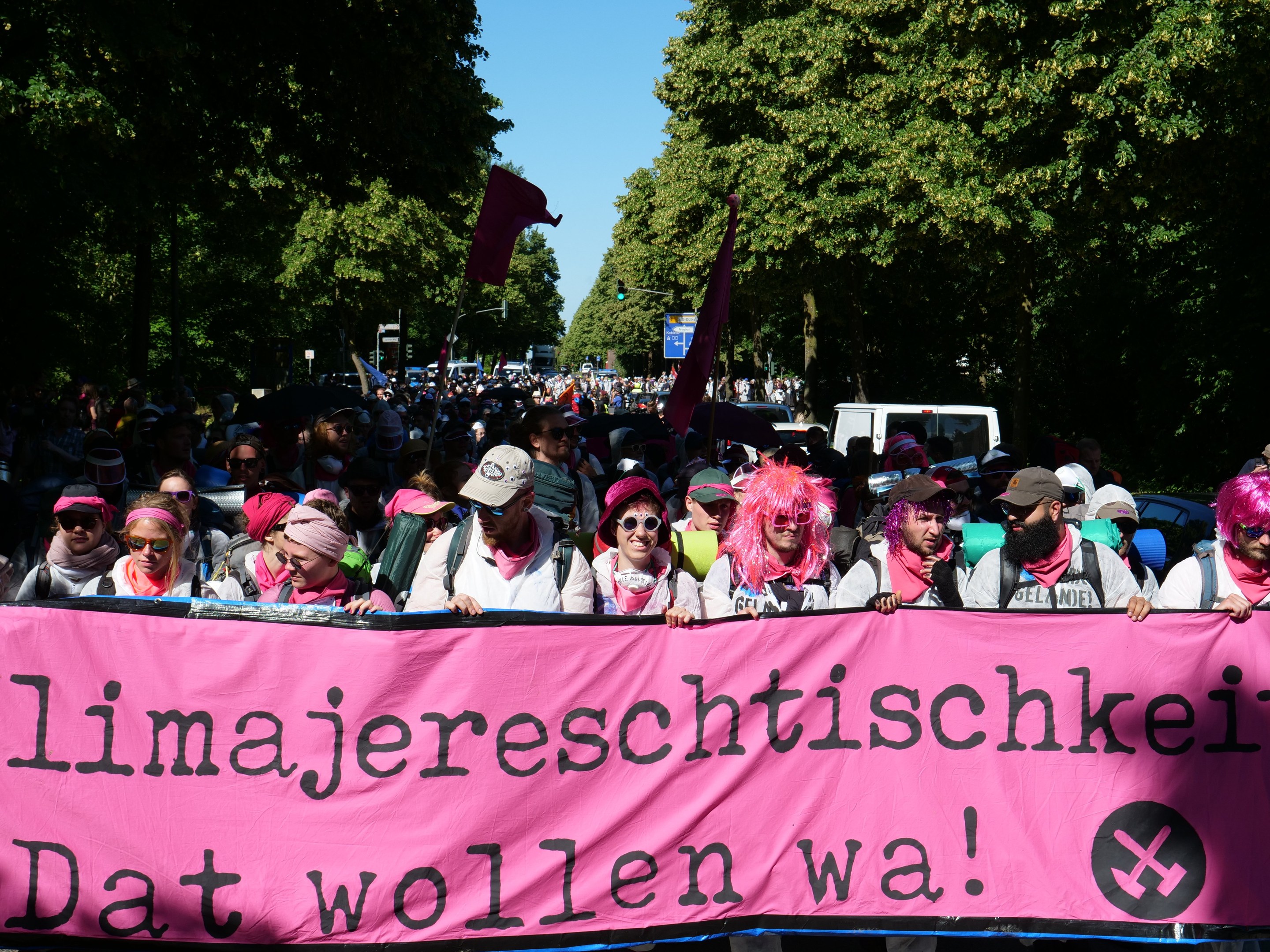 Eine Gruppe von Menschen marschiert mit einem pinken Banner, auf dem 'Frauenmarsch in Berlin' steht, durch die Straße. Einige tragen Mötzen und Schutzbrillen, andere halten Fahnen. Im Hintergrund sind Bäume, Fahrzeuge und der Himmel zu sehen.