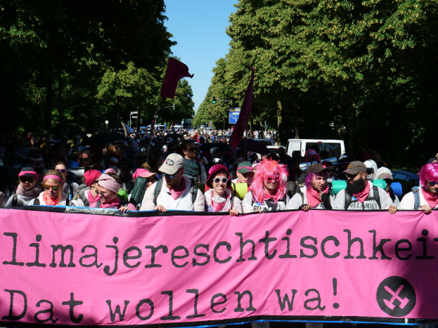 Eine Gruppe von Menschen marschiert mit einem pinken Banner, auf dem 'Frauenmarsch in Berlin' steht, durch die Straße. Einige tragen Mötzen und Schutzbrillen, andere halten Fahnen. Im Hintergrund sind Bäume, Fahrzeuge und der Himmel zu sehen.